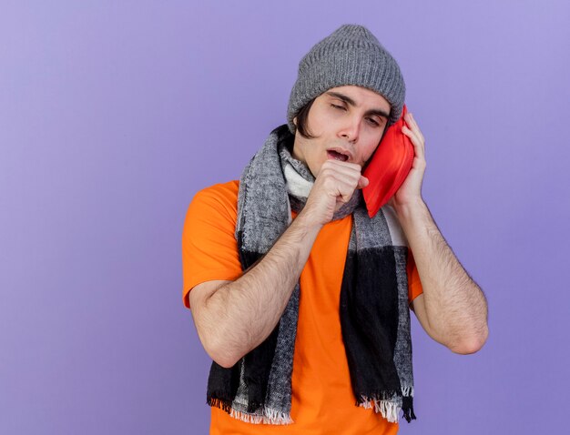 Toux jeune homme malade portant un chapeau d'hiver avec écharpe mettant le sac d'eau chaude sur la joue isolé sur fond violet