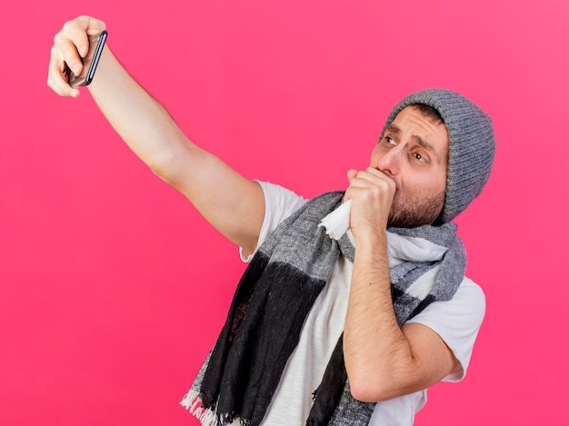 Photo gratuite toux jeune homme malade portant chapeau d'hiver avec écharpe prendre un selfie et mettre la main sur la bouche isolé sur fond rose