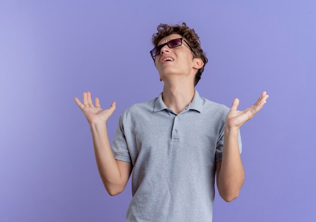 Tracassé jeune homme à lunettes noires portant un polo gris levant la main avec une expression agacée sur bleu