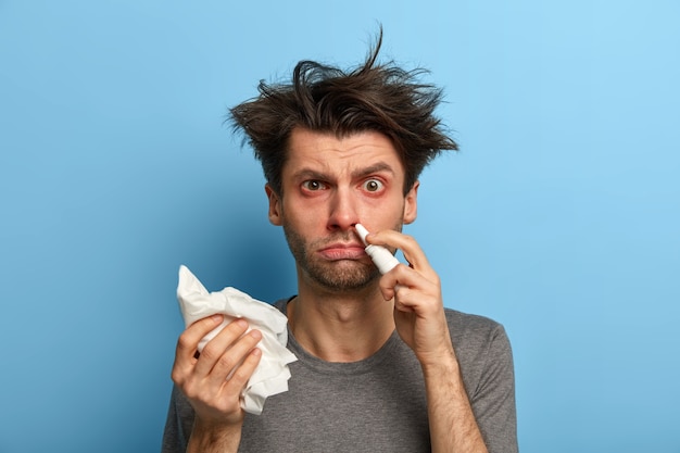 Traitement à domicile, virus, maladie saisonnière et concept d'allergie. Un homme mécontent casse le nez bouché, attrapé froid, tient un mouchoir, a de la fièvre, des yeux rouges et enflés, pose contre un mur bleu.