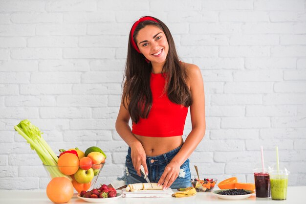 Tranche de découpage de femme souriante de banane sur planche à découper, debout contre le mur