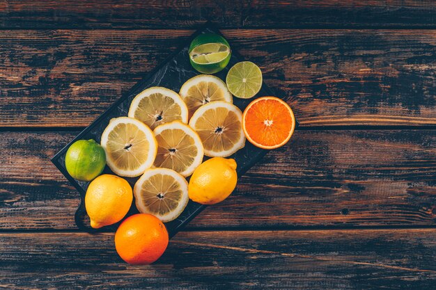 Tranches de citron avec des oranges et du citron vert à plat sur une planche à découper et un espace de fond en bois foncé pour le texte