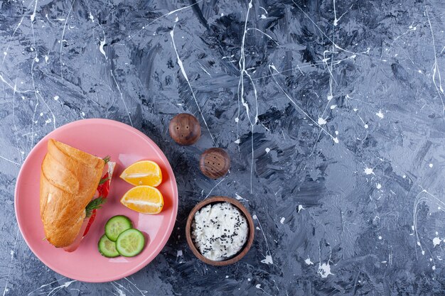 Tranches de citrons et de concombre, sandwich sur assiette à côté d'un bol de fromage sur bleu.