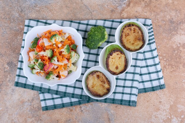 Tranches de courgettes frites sur un plateau, morceau de brocoli et un bol de salade de légumes sur une serviette sur une surface en marbre