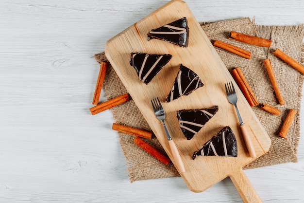 Tranches de gâteau au chocolat avec des bâtons de cannelle dans une planche de bois et un morceau de sac sur fond blanc, à plat.