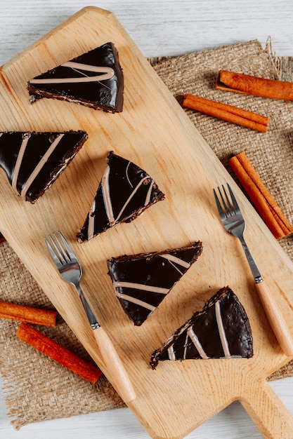 Photo gratuite tranches de gâteau au chocolat dans une planche de bois et morceau de sac avec des bâtons de cannelle et des fourchettes high angle view sur fond blanc