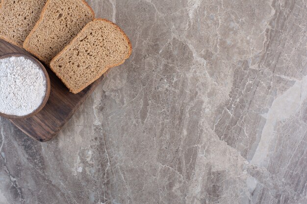 Tranches de pain noir et un bol de farine sur une planche en marbre.