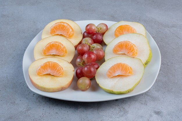 Tranches de pommes, poires, mandarines et raisins sur un plateau sur fond de marbre.