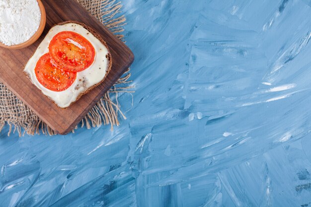 Tranches de tomates sur du pain au fromage et un bol de farine sur une planche à découper sur une serviette en toile de jute sur bleu.