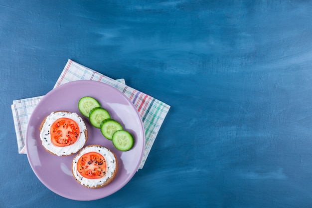 Tranches de tomates sur pain au fromage à côté de concombre sur plaque sur torchon, sur bleu.