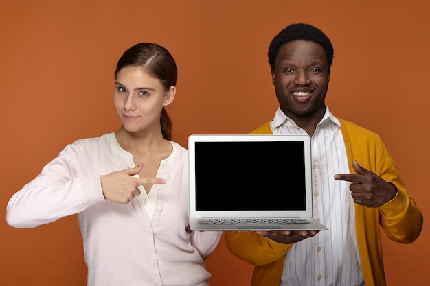 Travail d'équipe, gadgets électroniques et concept d'occupation. Heureux jeune femme européenne confiante et positif élégant mâle à la peau sombre pointant les doigts avant à l'écran d'ordinateur portable copyspace vierge et souriant