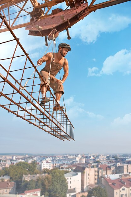 Travailler avec soin. Torse nu fort rétro constructeur regardant quelque chose attentivement debout sur une barre transversale suspendue à une grue low angle shot