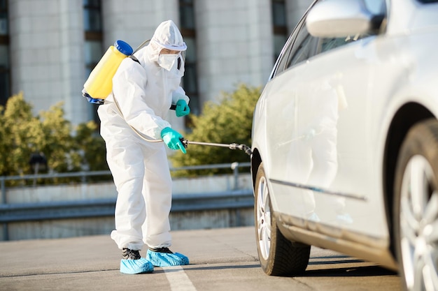 Travailleur de l'assainissement en tenue de protection pulvérisant un pneu de voiture pendant la désinfection de la ville en raison de la pandémie COVID19