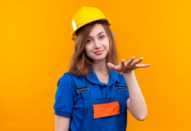 Travailleur de constructeur de jeune femme optimiste en uniforme de construction et casque de sécurité demandant se détendre, faire un geste de calme avec la main en souriant