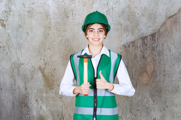 Travailleur de la construction féminin en casque vert tenant un marteau et donnant le pouce vers le haut