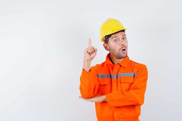 Travailleur de la construction pointant vers le haut en uniforme, casque et à la surprise, vue de face.