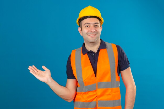 Travailleur de la construction portant un casque de sécurité blanc et un gilet, avec un sourire sur le visage pointant avec la paume de la main à l'espace de copie sur bleu isolé