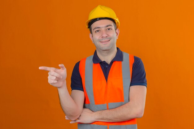 Travailleur de la construction portant un gilet et un casque de sécurité jaune pointant son index sur le côté avec un sourire sur le visage isolé sur orange