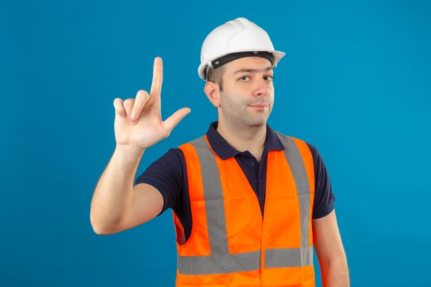 Travailleur de la construction portant un gilet et un casque de sécurité pointant avec le doigt vers le haut et l'expression en colère isolé sur bleu
