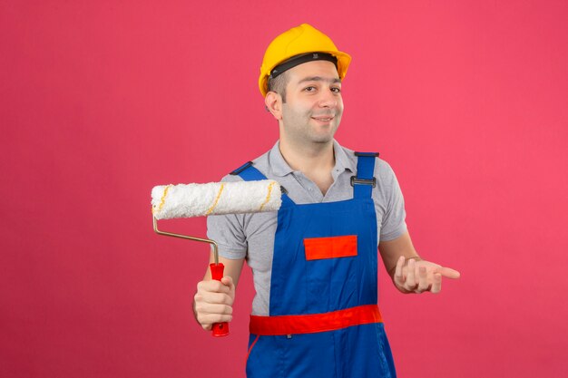 Travailleur de la construction portant un uniforme et un casque de sécurité faisant un geste confus avec la main et l'expression comme posant une question tenant un rouleau à peinture isolé sur rose
