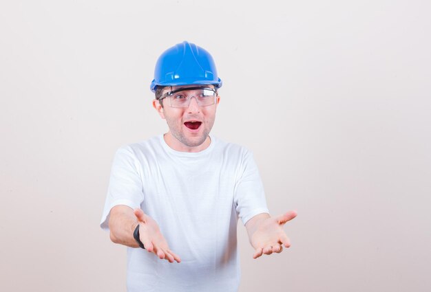 Travailleur de la construction s'étirant les mains de manière interrogative en t-shirt, casque et semblant étonné