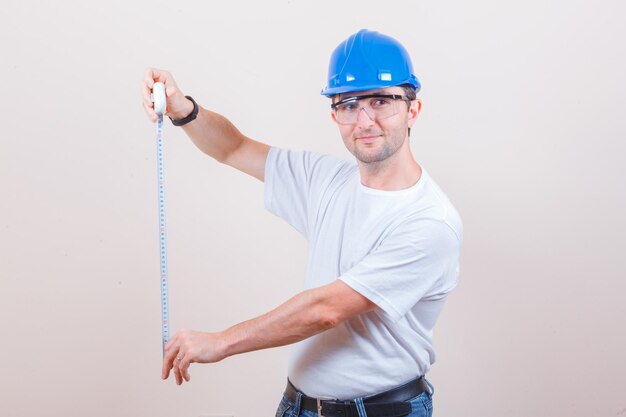 Travailleur de la construction en t-shirt, jeans, casque tenant un ruban à mesurer et regardant jolly