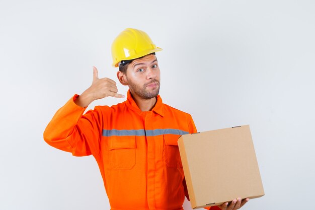 Travailleur de la construction tenant une boîte en carton, montrant le geste du téléphone en uniforme, casque et à la douce. vue de face.