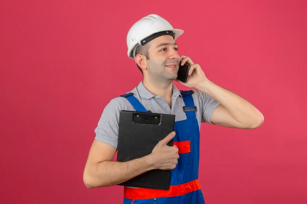 Travailleur de la construction en uniforme et casque de sécurité s'exprimant par téléphone mobile tenant le presse-papiers positif à la recherche isolé sur rose