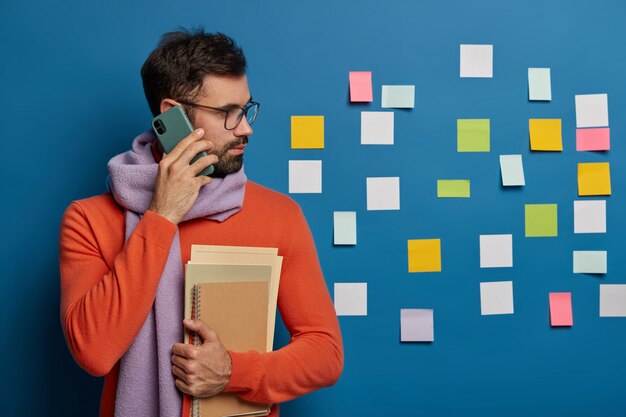 Travailleur créatif avec barbe appelle quelqu'un via téléphone portable, porte des lunettes, un pull avec une écharpe, regarde de côté sur des notes colorées collées sur un mur bleu