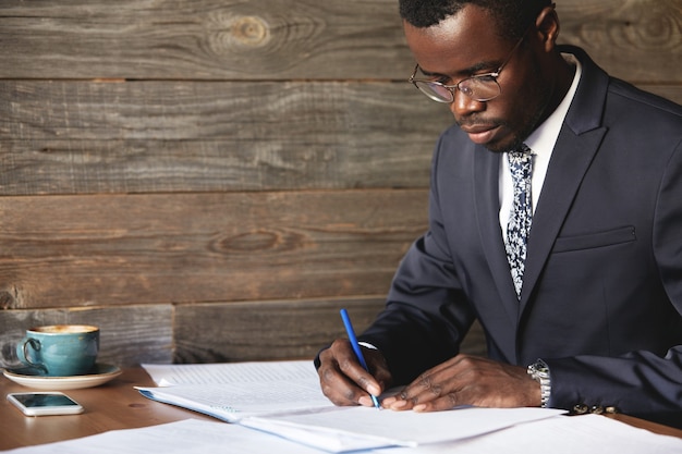 Travailleur d'entreprise noir sérieux en costume formel et lunettes signant un contrat lucratif