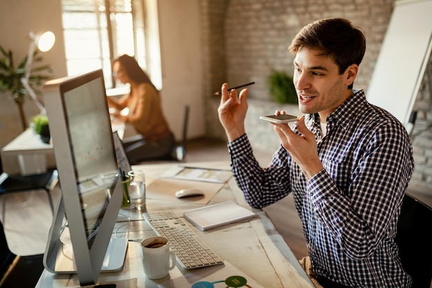 Photo gratuite travailleur indépendant souriant communiquant sur le haut-parleur du téléphone portable tout en lisant des rapports sur un ordinateur au bureau