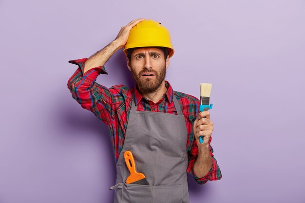 Travailleur industriel déçu vêtu d'un casque de sécurité, uniforme décontracté, tient un pinceau pour la peinture, étant peintre professionnel, a déplu l'expression du visage, isolé sur un mur violet