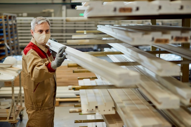 Travailleur masculin avec masque facial empilant des planches sur un rack à l'atelier de menuiserie