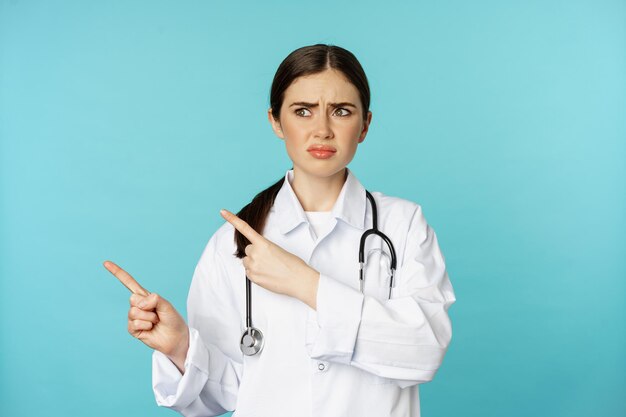 Travailleur médical confus et déçu, fronçant les sourcils tout en pointant les doigts vers la gauche avec une expression de visage douteuse et perplexe, debout sur fond de torquoise