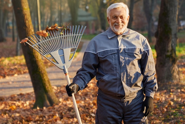 Photo gratuite travailleur senior souriant posant devant la caméra tout en rassemblant des feuilles d'automne en plein air portrait de heureux