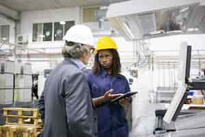 Photo gratuite travailleur d'usine féminin sérieux parlant au patron à la machine sur le sol de l'usine, pointant sur l'écran de la tablette