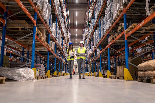 Les travailleurs avec des casques et des vestes réfléchissantes marchant dans une grande allée d'entrepôt vérifiant l'état des marchandises