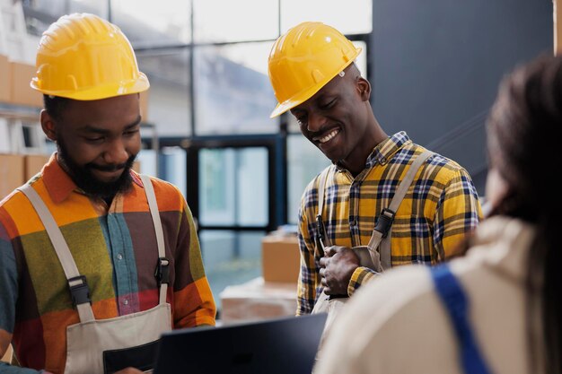 Des travailleurs d'entrepôt portant des casques jaunes rient et discutent au travail. Des hommes afro-américains souriants employés d'entrepôt dans des casques de protection et en général ayant une conversation amusante