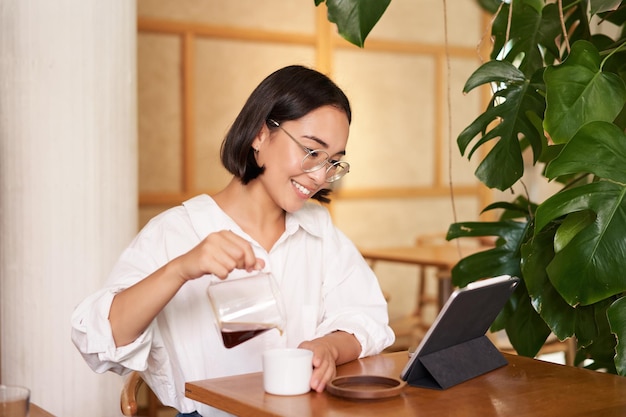 Travailleurs Indépendants Et à Distance Souriante Jeune Femme Versant Du Café Dans Une Tasse Assise Dans Un Café Et Regardant