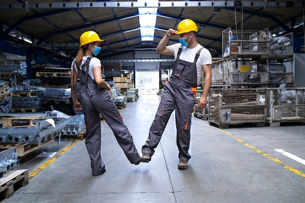 Travailleurs portant des uniformes et un casque à l'usine touchant avec les jambes et saluant en raison du virus corona et de l'infection