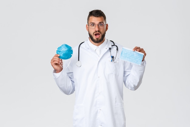 Travailleurs de la santé, assurance médicale, concept de pandémie et de covid-19. Médecin excité en lunettes et blouse blanche, médecin montrant un masque médical et un respirateur, haletant étonné