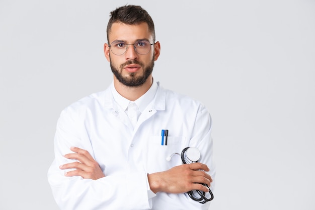 Travailleurs de la santé, coronavirus, pandémie de covid-19 et concept d'assurance. Gros plan sur un jeune médecin sérieux en blouse blanche, lunettes, écoute attentivement le patient, la poitrine des bras croisés, tenant un stéthoscope.