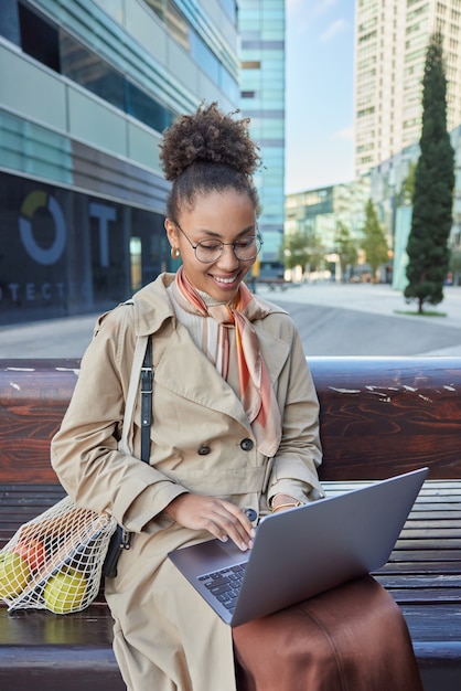 une travailleuse à distance travaille des claviers indépendants sur un ordinateur portable assise sur un banc en bois à l'extérieur porte des lunettes rondes et un manteau porte un sac en filet travaille sur le projet