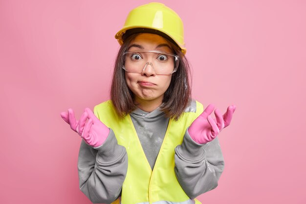 Une travailleuse industrielle asiatique hésitante et perplexe écarte les paumes de ses regards avec une expression désemparée ne sait pas par quoi commencer à travailler sur un chantier de construction vêtue d'un uniforme