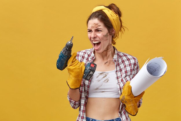 Travailleuse industrielle en colère tenant du papier roulé et une perceuse en la regardant et en hurlant d'être furieuse à cause de la panne de son instrument. Travailleuse émotionnelle ayant des problèmes pendant le travail