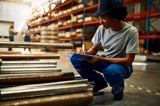 Travailleuse noire écrivant sur le presse-papiers lors de l'inspection de produits en acier dans un entrepôt industriel
