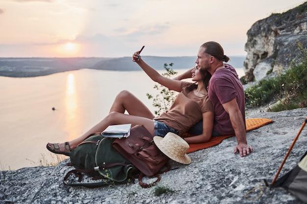 Étreindre couple avec sac à dos assis au sommet de la montagne rocheuse bénéficiant d'une vue sur la côte d'une rivière ou d'un lac.