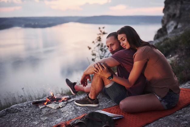 Étreindre le couple avec sac à dos assis près du feu au sommet de la montagne en profitant de la vue sur la côte d'une rivière ou d'un lac.