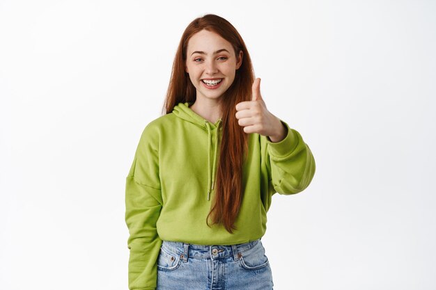 Très bien. Une adolescente rousse souriante montre les pouces vers le haut et a l'air satisfaite, loue beaucoup, recommande un magasin, fait un compliment, fond blanc.