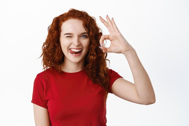 Très bonne fille au gingembre souriante montrant un signe correct et un clin d'œil à la caméra dire oui approuver quelque chose de bien comme et accepter louange excellent choix fond blanc